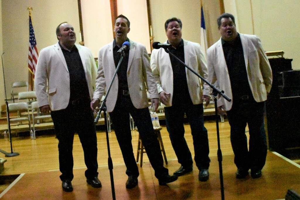 4 men in white jackets, singing.